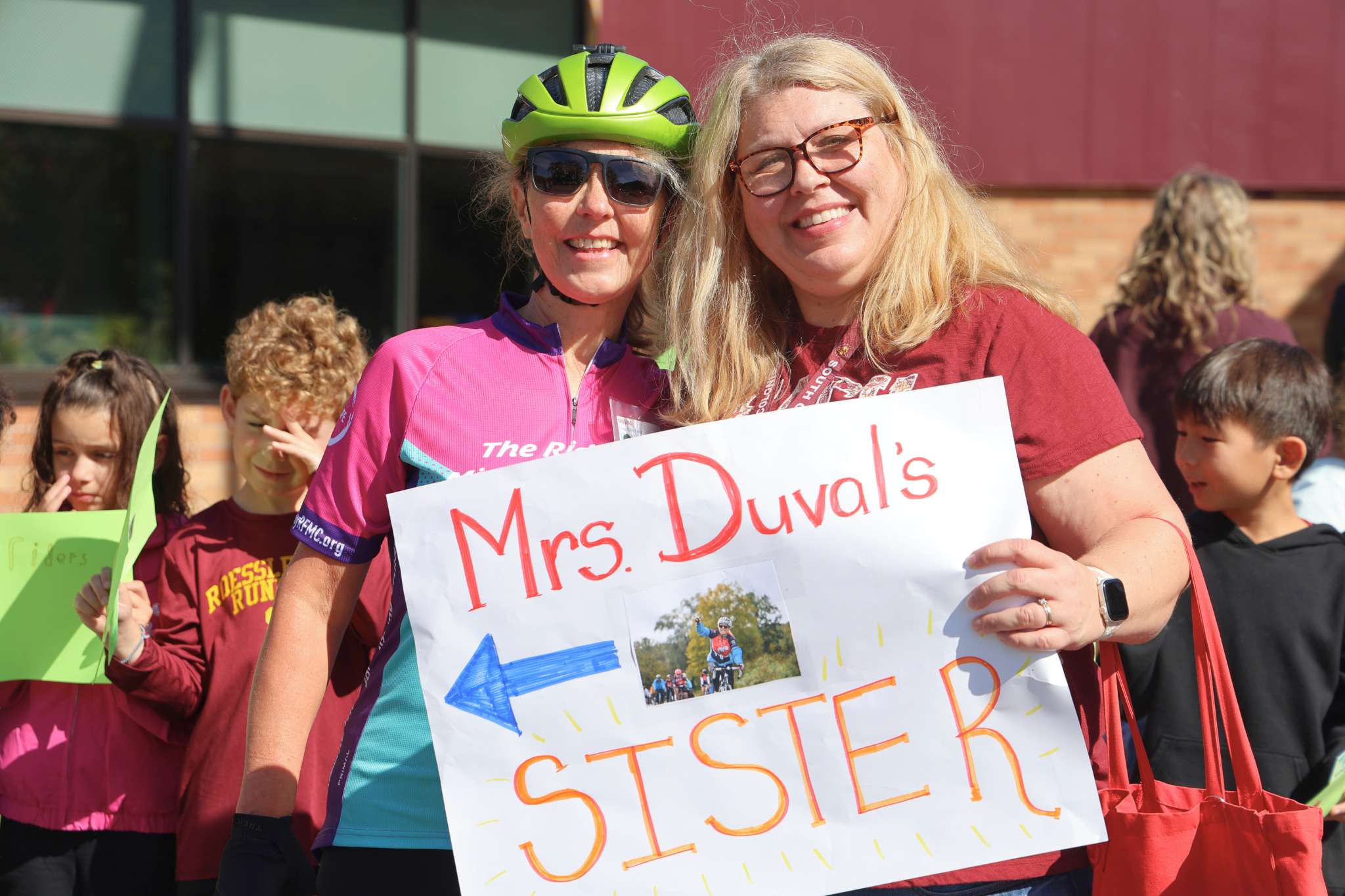 Two women smile while holding a sign that reads 'Mrs Duval's SISTER' with an arrow.